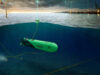 Green autonomous underwater vehicles tethered by cables on a seabed near an offshore wind farm and port at dusk, with the surface showing wind turbines and lights.