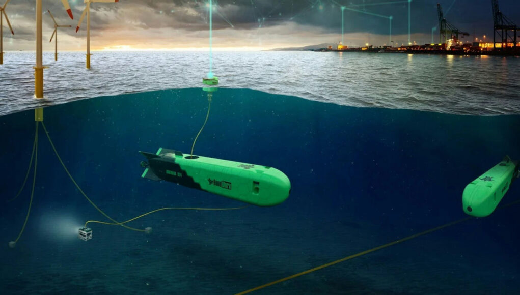 Green autonomous underwater vehicles tethered by cables on a seabed near an offshore wind farm and port at dusk, with the surface showing wind turbines and lights.