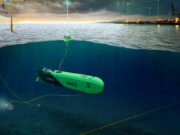 Green autonomous underwater vehicles tethered by cables on a seabed near an offshore wind farm and port at dusk, with the surface showing wind turbines and lights.
