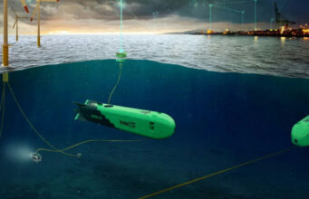 Green autonomous underwater vehicles tethered by cables on a seabed near an offshore wind farm and port at dusk, with the surface showing wind turbines and lights.