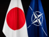 Two flags&mdash;Japan (white with red circle) and NATO (blue with white star-like emblem)&mdash;hung side by side against a dark background, symbolizing international cooperation.
