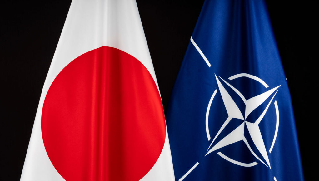 Two flags&mdash;Japan (white with red circle) and NATO (blue with white star-like emblem)&mdash;hung side by side against a dark background, symbolizing international cooperation.