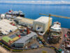 Aerial view of a busy shipyard by the sea with large white hangars, a yellow crane, and ships docked along the water's edge, surrounded by vehicles and buildings.