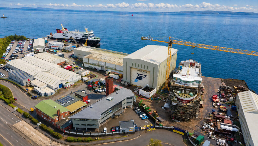 Aerial view of a busy shipyard by the sea with large white hangars, a yellow crane, and ships docked along the water's edge, surrounded by vehicles and buildings.