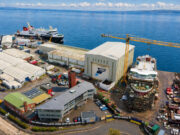 Aerial view of a busy shipyard by the sea with large white hangars, a yellow crane, and ships docked along the water's edge, surrounded by vehicles and buildings.