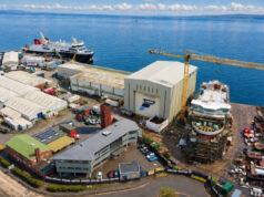 Aerial view of a busy shipyard by the sea with large white hangars, a yellow crane, and ships docked along the water's edge, surrounded by vehicles and buildings.