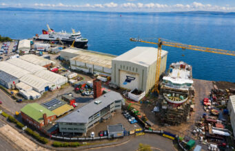 Aerial view of a busy shipyard by the sea with large white hangars, a yellow crane, and ships docked along the water's edge, surrounded by vehicles and buildings.