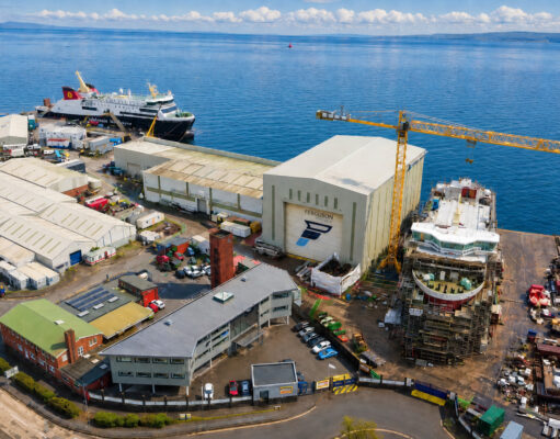 Aerial view of a busy shipyard by the sea with large white hangars, a yellow crane, and ships docked along the water's edge, surrounded by vehicles and buildings.