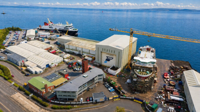 Aerial view of a busy shipyard by the sea with large white hangars, a yellow crane, and ships docked along the water's edge, surrounded by vehicles and buildings.