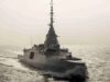 Surface warship sailing on calm sea with wake trailing behind, sun glare on the water.