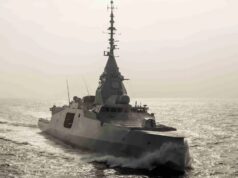 Surface warship sailing on calm sea with wake trailing behind, sun glare on the water.