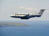 Twin-turboprop Royal Navy aircraft (ZZ503) in white and blue livery, flying low over the coastline and sea.