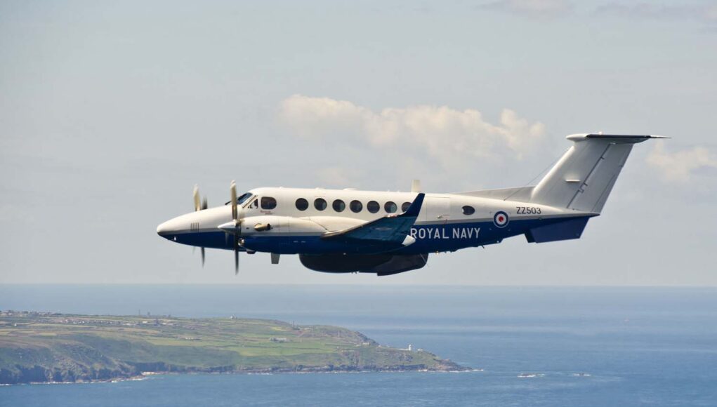 Twin-turboprop Royal Navy aircraft (ZZ503) in white and blue livery, flying low over the coastline and sea.