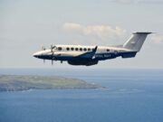 Twin-turboprop Royal Navy aircraft (ZZ503) in white and blue livery, flying low over the coastline and sea.