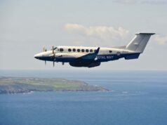 Twin-turboprop Royal Navy aircraft (ZZ503) in white and blue livery, flying low over the coastline and sea.