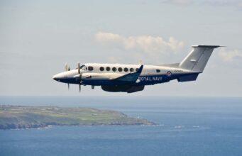 Second upgraded Avenger takes flight at Culdrose Twin-turboprop Royal Navy aircraft (ZZ503) in white and blue livery, flying low over the coastline and sea.