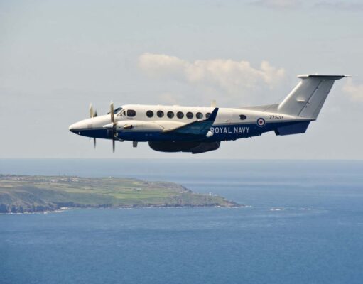 Twin-turboprop Royal Navy aircraft (ZZ503) in white and blue livery, flying low over the coastline and sea.