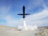 Black jet lifting off from a sandy desert airstrip, exhaust plume blasting upward and creating a cloud at ground level.