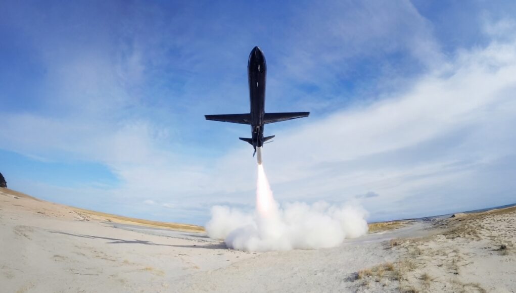 Black jet lifting off from a sandy desert airstrip, exhaust plume blasting upward and creating a cloud at ground level.