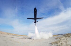 Black jet lifting off from a sandy desert airstrip, exhaust plume blasting upward and creating a cloud at ground level.