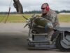 Airman in camouflage with red ear protection inspecting a large green missile on a ground launch trolley on an airfield.