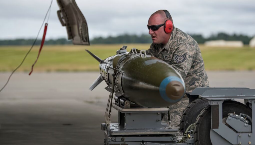 Airman in camouflage with red ear protection inspecting a large green missile on a ground launch trolley on an airfield.