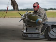 Airman in camouflage with red ear protection inspecting a large green missile on a ground launch trolley on an airfield.