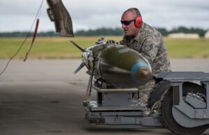 Airman in camouflage with red ear protection inspecting a large green missile on a ground launch trolley on an airfield.