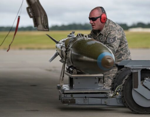 Airman in camouflage with red ear protection inspecting a large green missile on a ground launch trolley on an airfield.