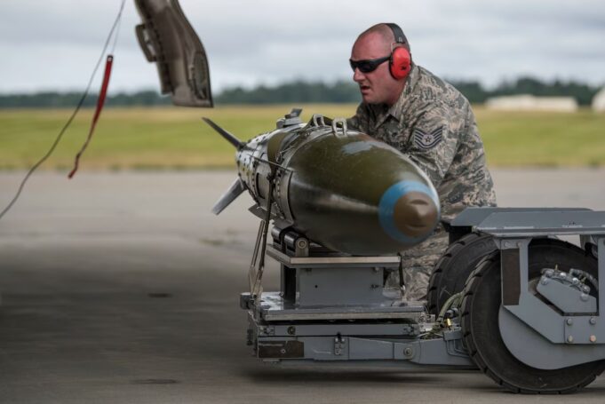 Airman in camouflage with red ear protection inspecting a large green missile on a ground launch trolley on an airfield.