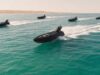 Five black speedboats race across turquoise water, leaving white wake with a sandy desert coastline in the background.