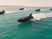 Britain sending a drone fleet to Hormuz Five black speedboats race across turquoise water, leaving white wake with a sandy desert coastline in the background.