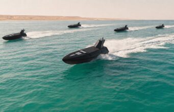 Britain sending a drone fleet to Hormuz Five black speedboats race across turquoise water, leaving white wake with a sandy desert coastline in the background.