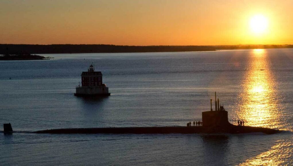 Sunset over calm water with a long submarine-like vessel and a small platform, silhouettes of people on deck.