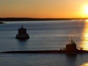 Sunset over calm water with a long submarine-like vessel and a small platform, silhouettes of people on deck.