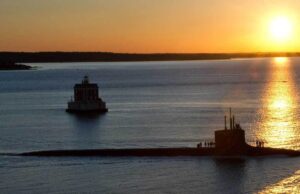 Sunset over calm water with a long submarine-like vessel and a small platform, silhouettes of people on deck.