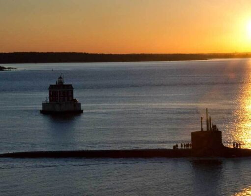 Sunset over calm water with a long submarine-like vessel and a small platform, silhouettes of people on deck.