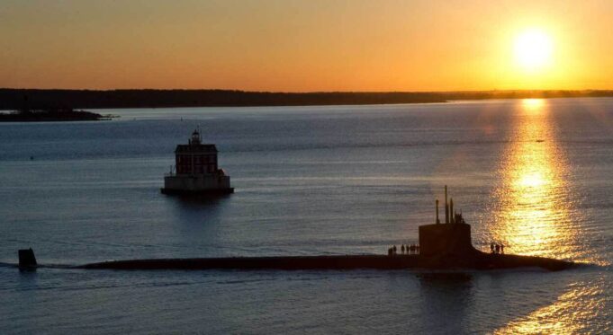 Sunset over calm water with a long submarine-like vessel and a small platform, silhouettes of people on deck.