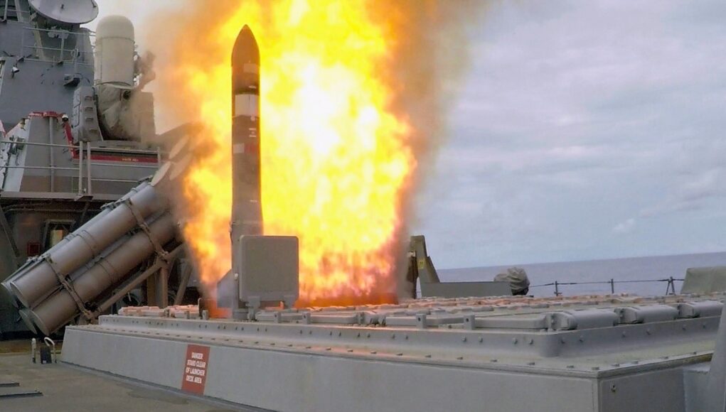 Missile launches from a naval ship's launcher with a large orange flame plume surrounding the rocket.