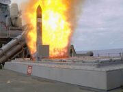 Missile launches from a naval ship's launcher with a large orange flame plume surrounding the rocket.