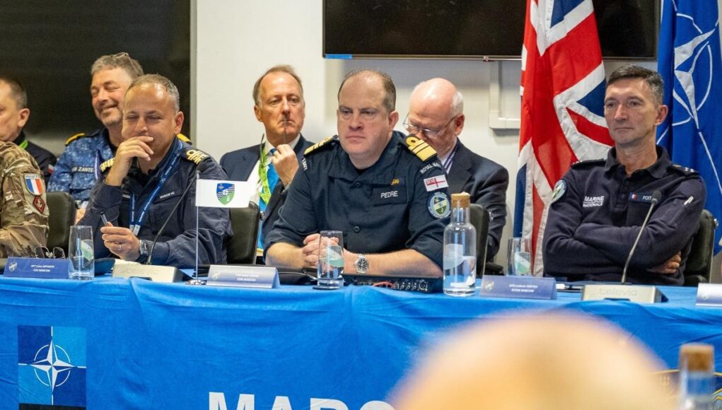 Panel of uniformed military officers seated at a blue NATO table with flags, microphones, and water bottles during a conference briefing.