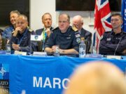 Panel of uniformed military officers seated at a blue NATO table with flags, microphones, and water bottles during a conference briefing.
