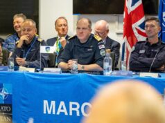 Panel of uniformed military officers seated at a blue NATO table with flags, microphones, and water bottles during a conference briefing.