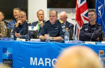 Panel of uniformed military officers seated at a blue NATO table with flags, microphones, and water bottles during a conference briefing.