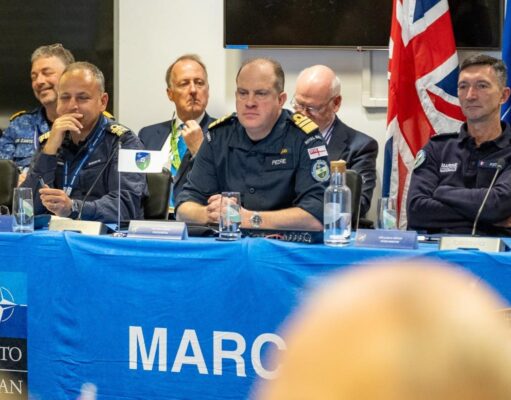 Panel of uniformed military officers seated at a blue NATO table with flags, microphones, and water bottles during a conference briefing.