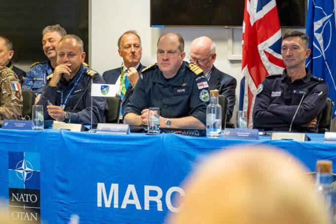 Panel of uniformed military officers seated at a blue NATO table with flags, microphones, and water bottles during a conference briefing.