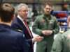 Man in a navy suit and red tie speaks to a group of uniformed pilots in a hangar-like setting, gesturing with his hands.