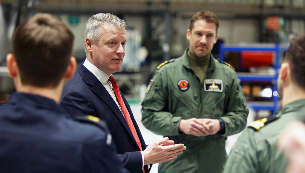 Man in a navy suit and red tie speaks to a group of uniformed pilots in a hangar-like setting, gesturing with his hands.