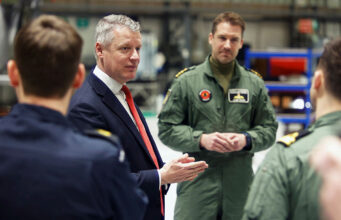 Man in a navy suit and red tie speaks to a group of uniformed pilots in a hangar-like setting, gesturing with his hands.