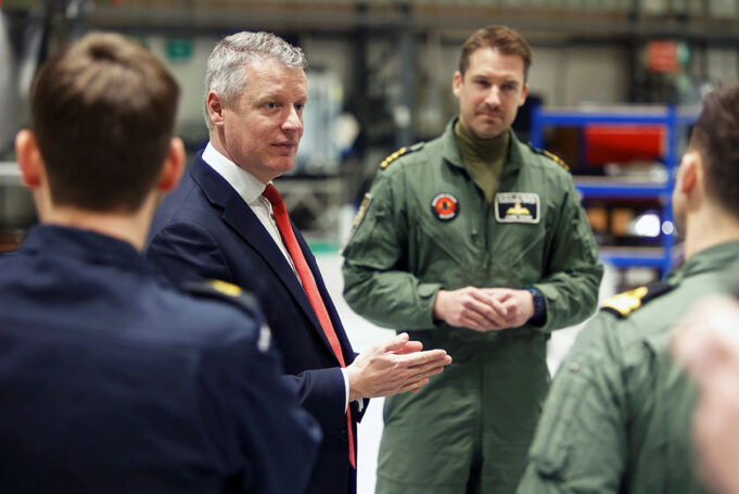 Man in a navy suit and red tie speaks to a group of uniformed pilots in a hangar-like setting, gesturing with his hands.